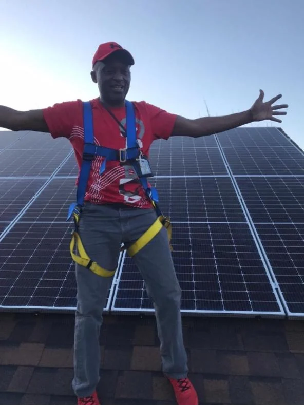An iThandimbvelo team member in a fall-protection harness in front of a rooftop array